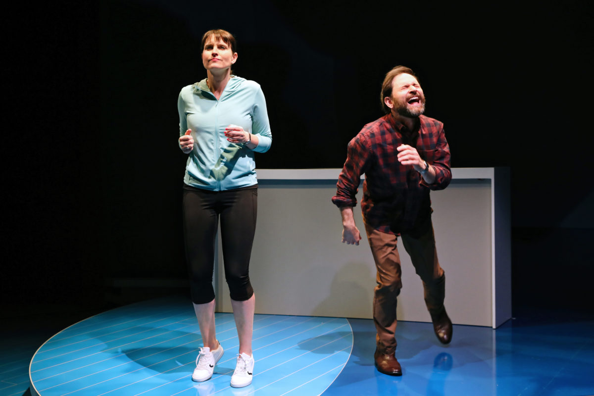 A woman in sportswear stands tensely with her fists clenched, while a bearded man in a checked shirt and brown trousers leans forward laughing, both on a blue circular stage with a white structure behind them.