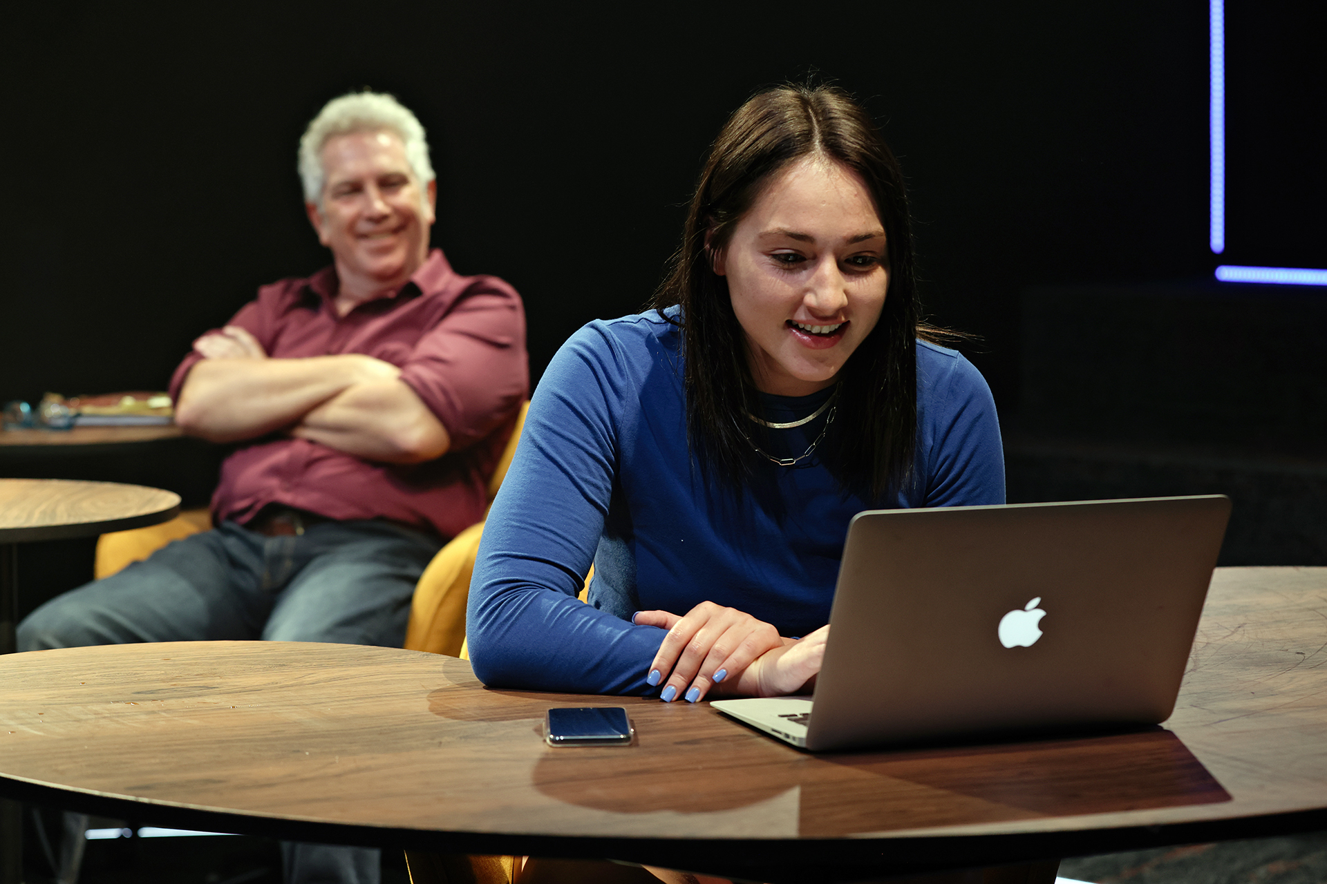 A young woman sits at a round table, smiling whilst using a laptop. A phone lies nearby. In the background, a man with grey hair and a maroon shirt sits with arms crossed, smiling and watching her.