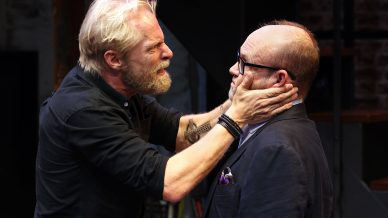 A bearded man urgently holds another man's face with both hands, speaking intensely, whilst the second man in glasses and a suit jacket looks uncomfortable. The scene suggests a dramatic moment on stage.