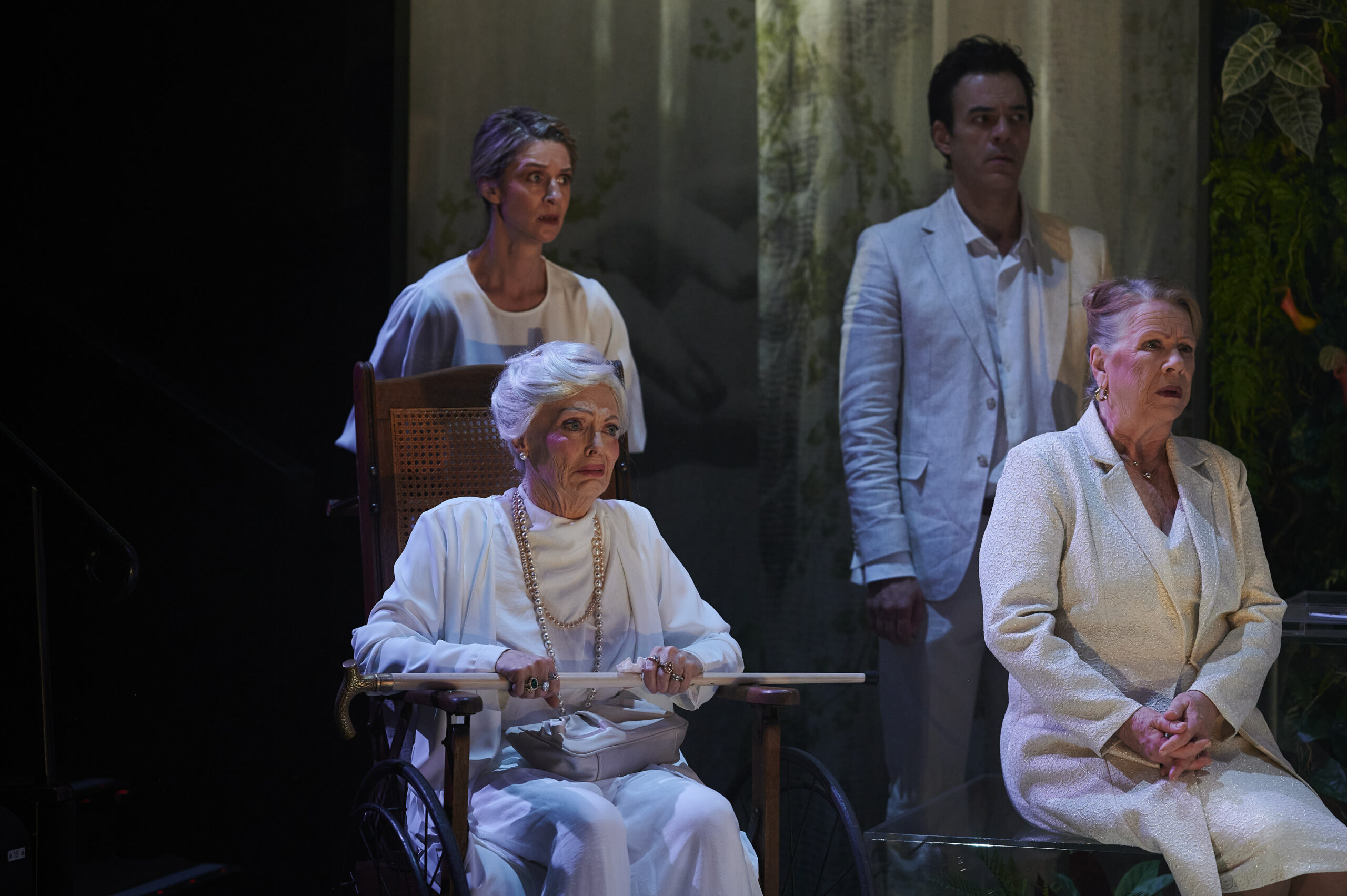 Four people dressed in white, three standing and one seated in a wheelchair, appear serious on a dimly lit stage with plants and sheer curtains in the background.