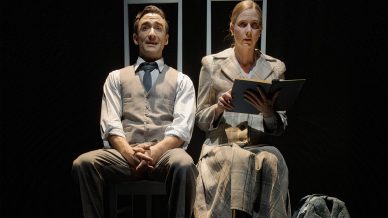A man and a woman sit side by side on chairs, dressed in formal attire. The woman reads from a folder whilst the man smiles. Their reflections are visible on the shiny black floor beneath them.