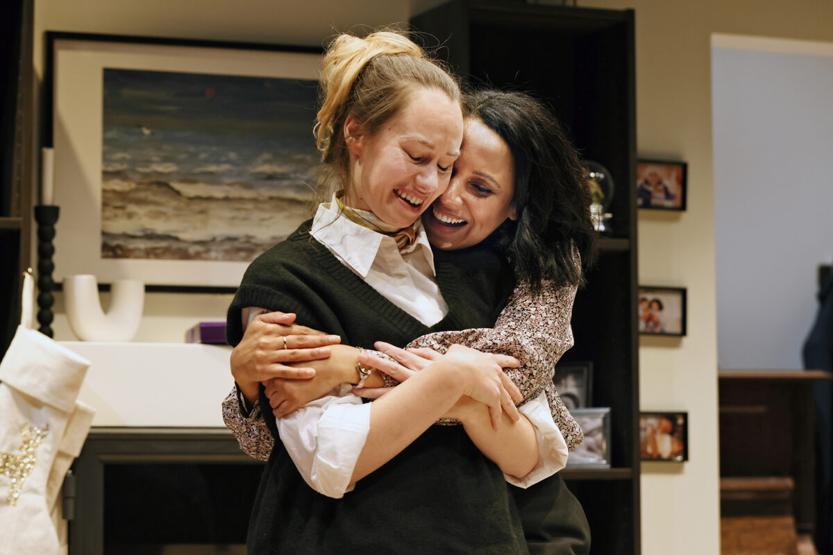Two women stand indoors embracing and smiling warmly. One woman closes her eyes and laughs whilst the other hugs her from behind with a joyful expression. Shelves and framed photos are visible in the background.