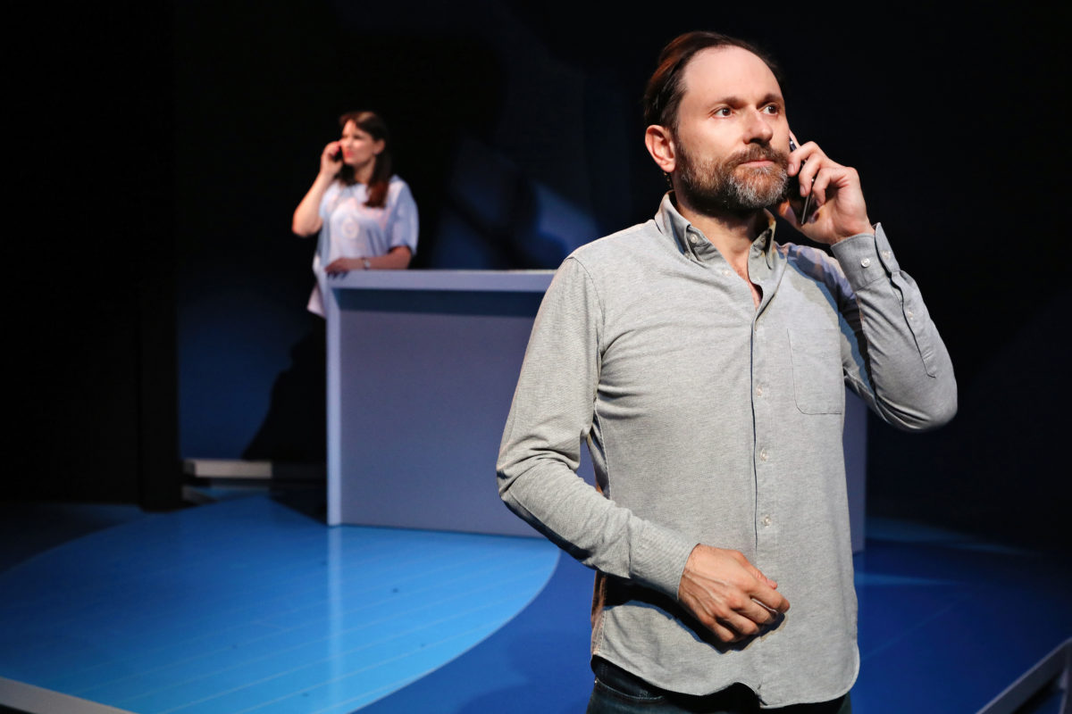 A bearded man in a grey shirt talks on his phone in the foreground, whilst a woman in a light blouse stands behind a counter in the background, also on the phone, on a stage with blue lighting.
