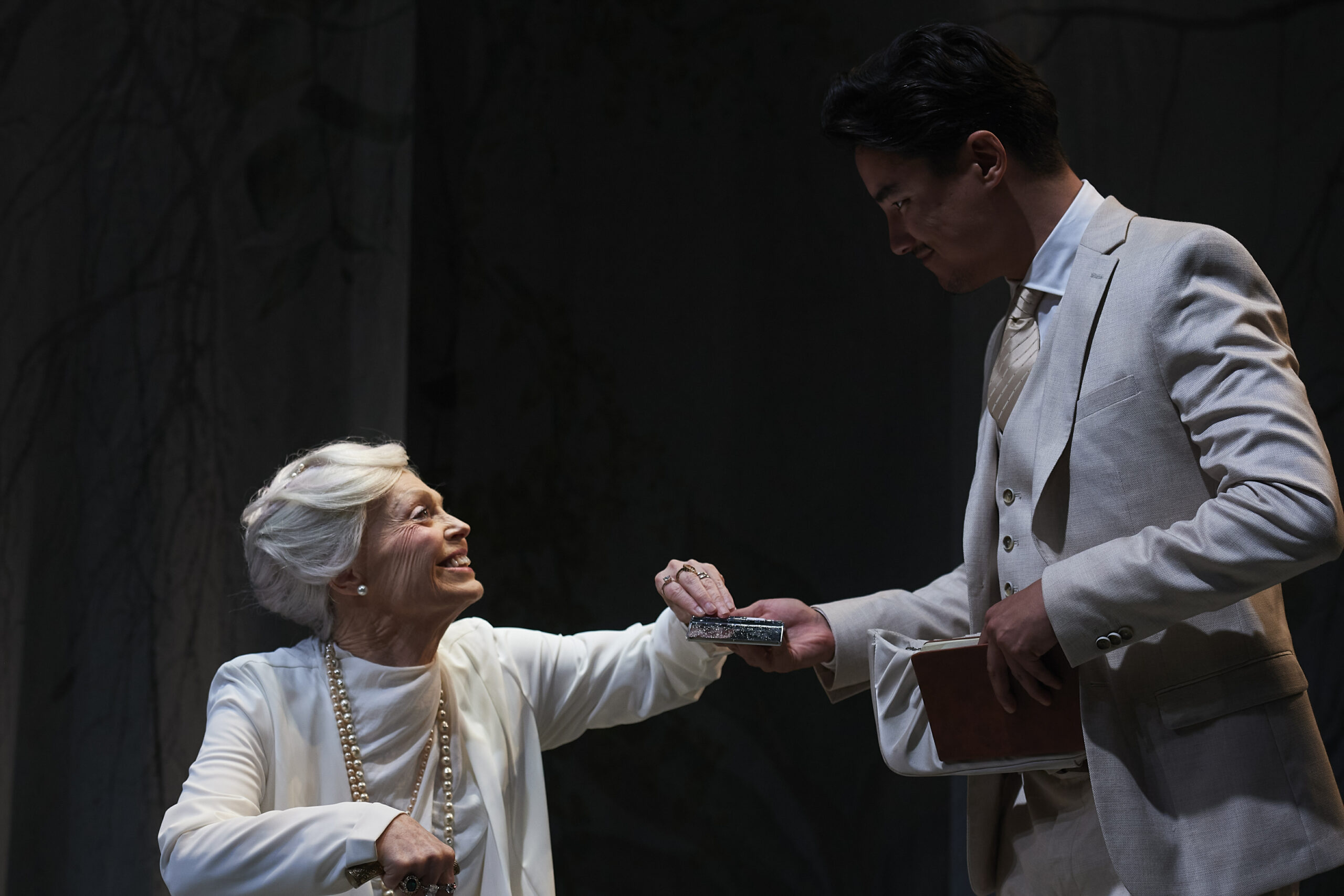 An older woman dressed in white smiles and reaches out to hand a small object to a younger man in a light-coloured suit who is holding a book. The scene is dimly lit, suggesting a theatrical performance.