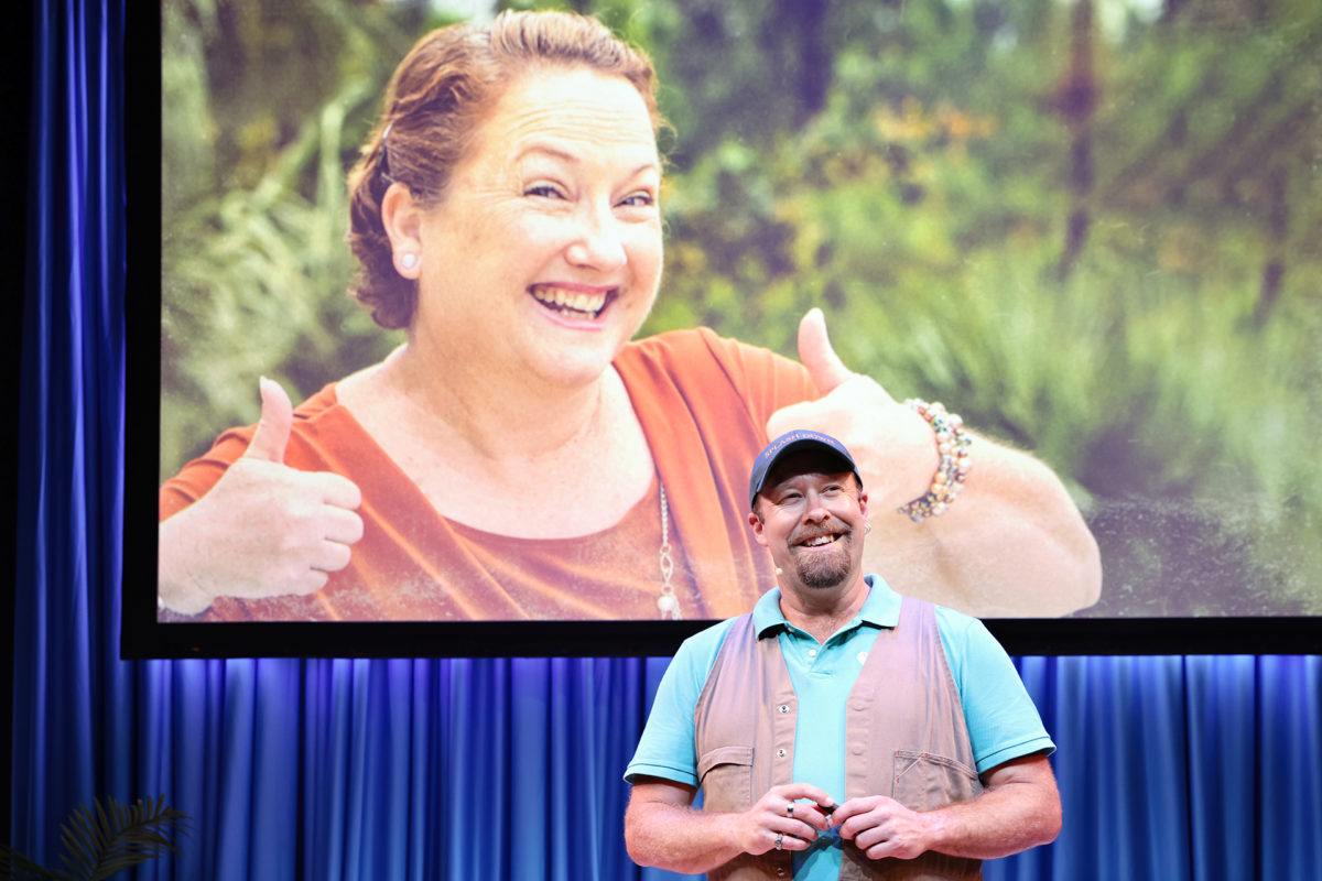 A man in a blue shirt and cap stands on stage, smiling, with a large screen behind him displaying a woman in an orange shirt giving two thumbs up and smiling broadly. Blue curtains form the stage backdrop.