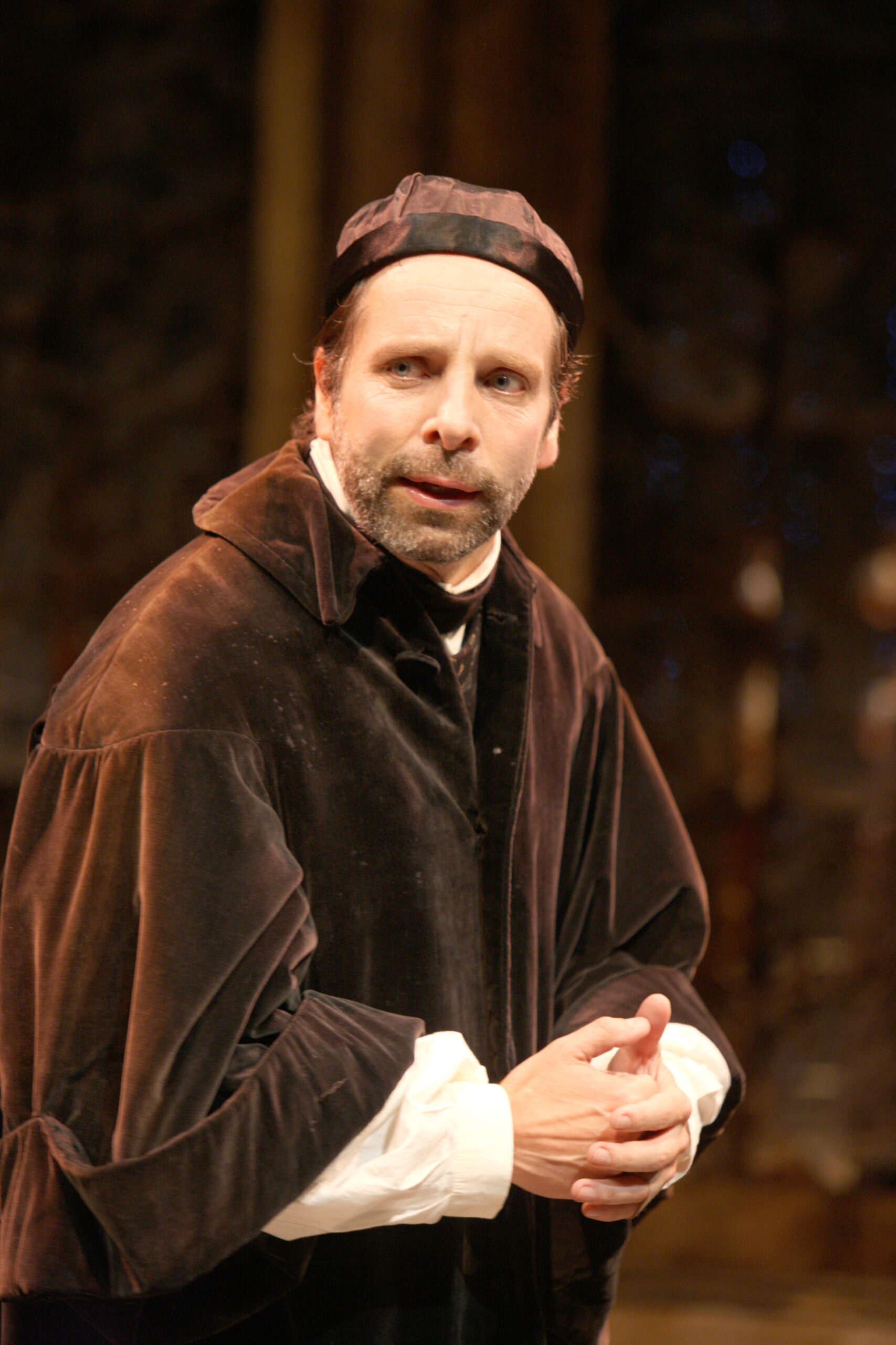 A man in historical clothing, including a black cap and dark velvet robe, stands with hands clasped, looking intently ahead, as if performing on stage in a dramatic scene.