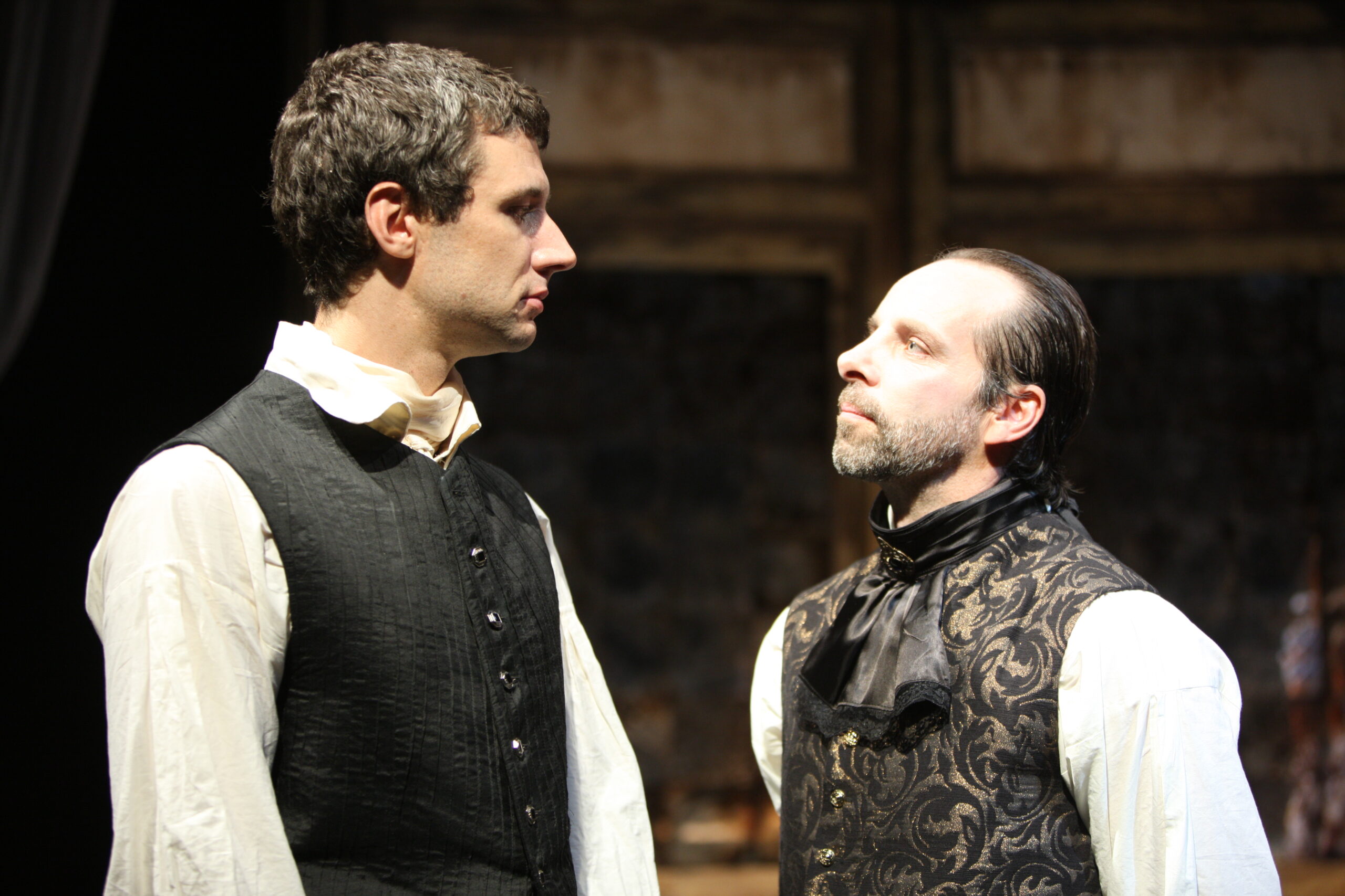 Two men in historical clothing stand facing each other intensely on stage, one looking at the other with a stern expression. The background is dimly lit with wooden panelling, suggesting a dramatic theatre scene.