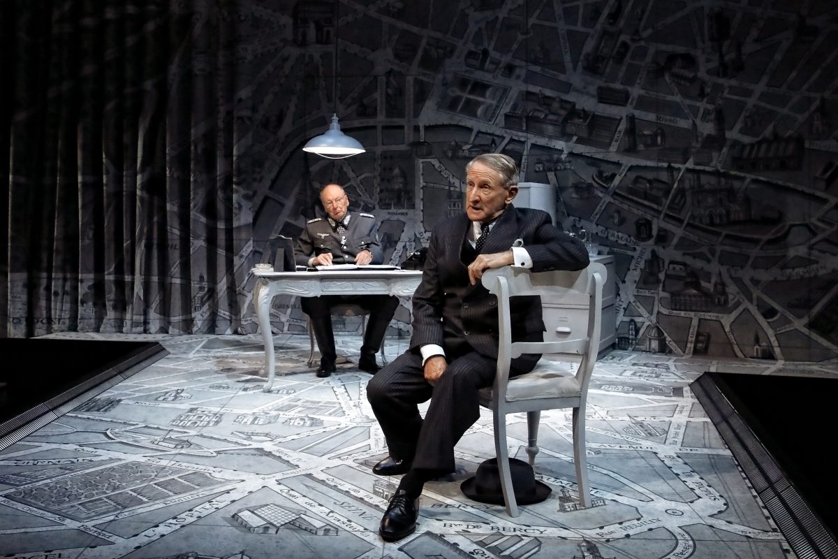 Two men in suits sit on a stage set designed like a map, one at a desk under a lamp writing, the other in front, seated sideways and looking thoughtful. The scene has a dramatic, theatrical atmosphere.
