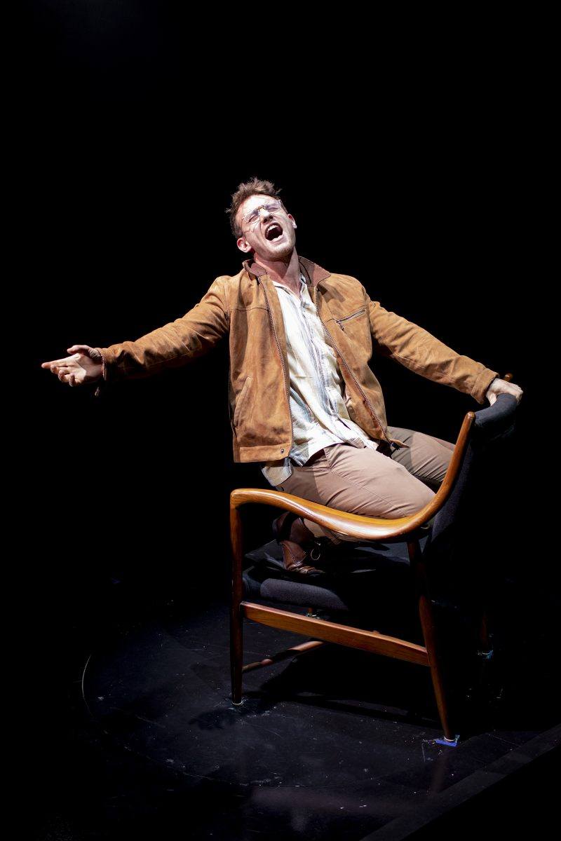 A man in a brown jacket and glasses sits backwards on a wooden chair, leaning back and screaming or singing passionately, with one arm outstretched, on a dark stage under dramatic lighting.