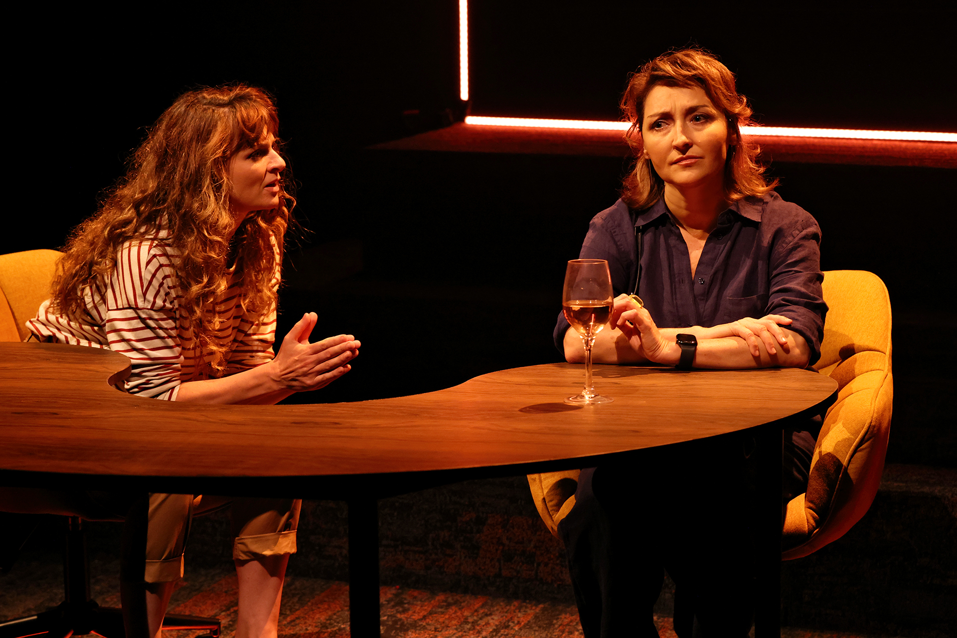 Two women sit at a curved wooden table under warm lighting. One woman gestures whilst speaking, looking engaged; the other looks thoughtful, resting her arm on the table beside a glass of wine.