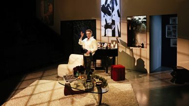 A person stands on a modern, warmly lit living room set with a white sofa, glass coffee table, piano, and photographs. Shadows and art decorate the walls, creating a dramatic ambience.