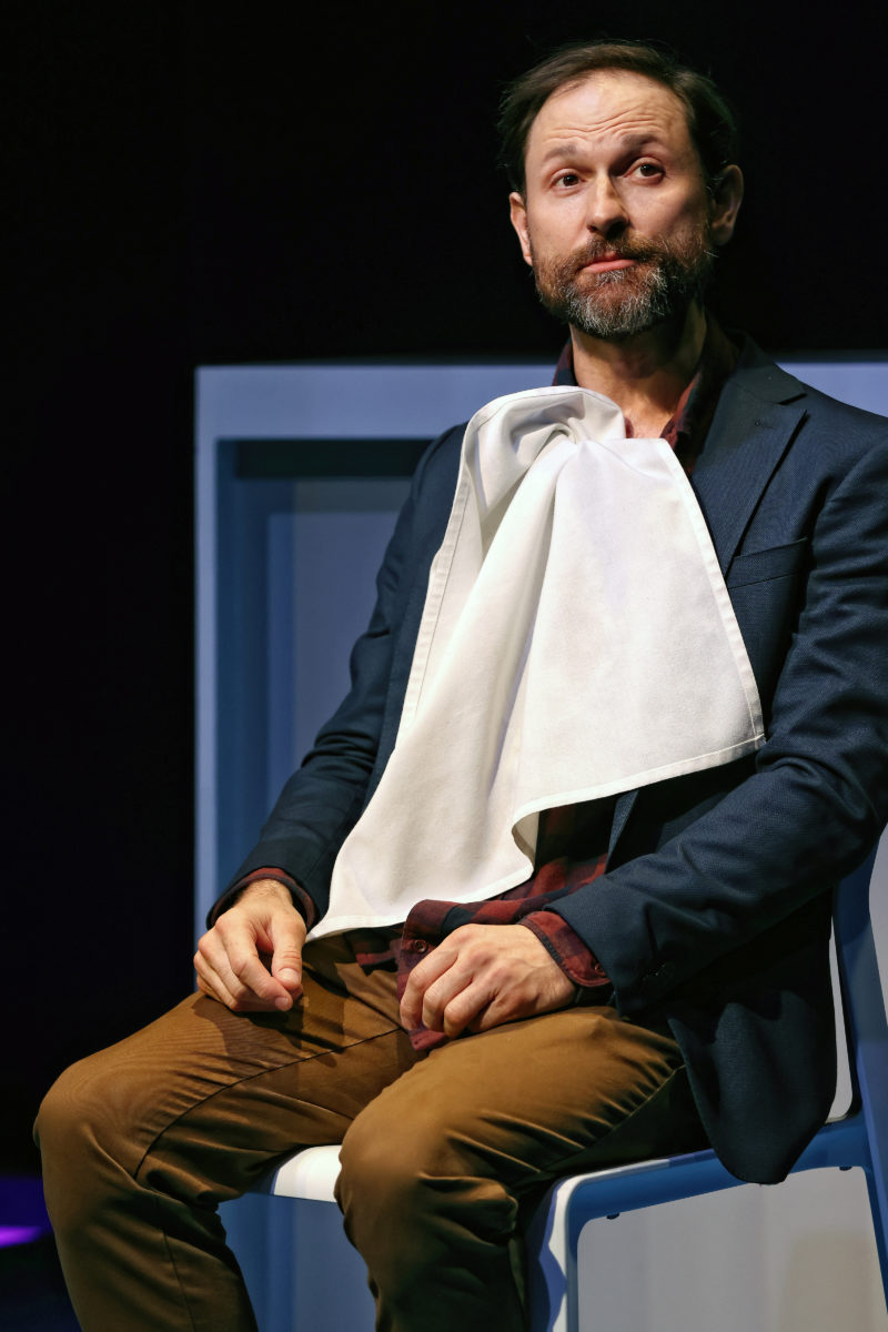 A man with a beard sits on a chair, wearing a dark blazer and brown trousers, with a white serviette tucked into his collar. He looks to the side with a thoughtful expression against a dark background.