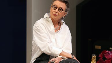 An older woman with short grey hair and round glasses sits holding a glass of white wine, wearing a white shirt, dark trousers, and a necklace, looking thoughtfully to the side. A champagne bottle and red flowers are nearby.