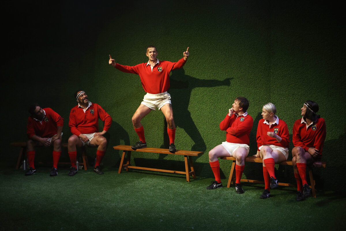 A man in a red rugby kit stands on a bench with arms raised, enthusiastically addressing four teammates who are seated and listening, against a green textured backdrop.