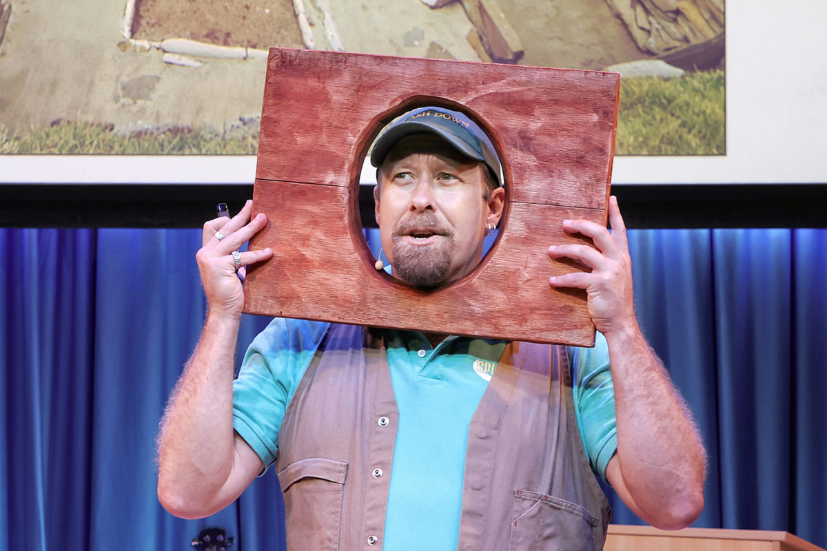 A man wearing a cap and waistcoat holds a wooden board with a circular hole around his face, standing on a stage with blue curtains and a projected image behind him.