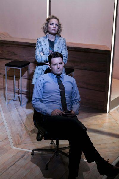 A woman stands with arms crossed behind a man sitting on a swivel chair. The man is dressed in a blue shirt and tie, holding a mobile. They are in a modern room with wooden furniture and minimalist decor.