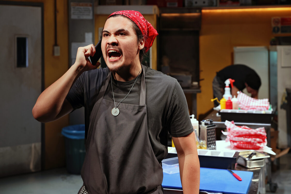 A man wearing a red bandanna and apron angrily speaks on the phone in a commercial kitchen, whilst another person works in the background. Various kitchen supplies and ingredients are visible around them.