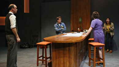 Four people stand around a wooden table in a dimly lit room, appearing to be in a heated discussion or argument. Two women face two men; one woman gestures emotionally while the others look tense or attentive.