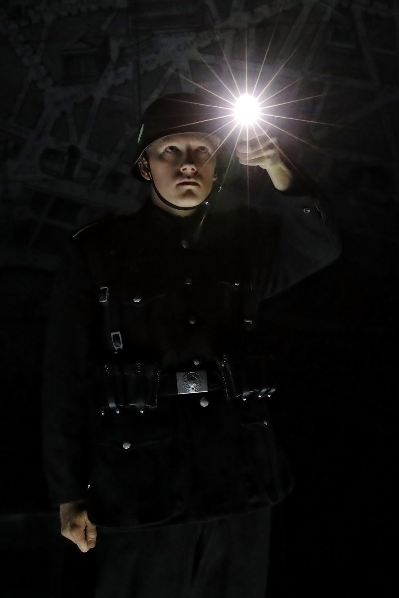 A person in a dark uniform and helmet holds a bright torch, illuminating their face in an otherwise dark setting. The background appears shadowy and indistinct.