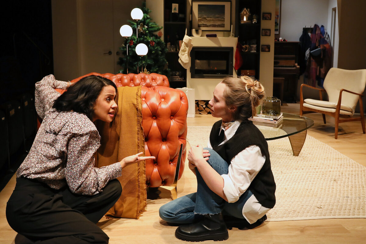Two women kneel on the floor beside an orange tufted sofa in a cosy, modern living room, engaged in a serious conversation. A decorated Christmas tree and various home furnishings are visible in the background.