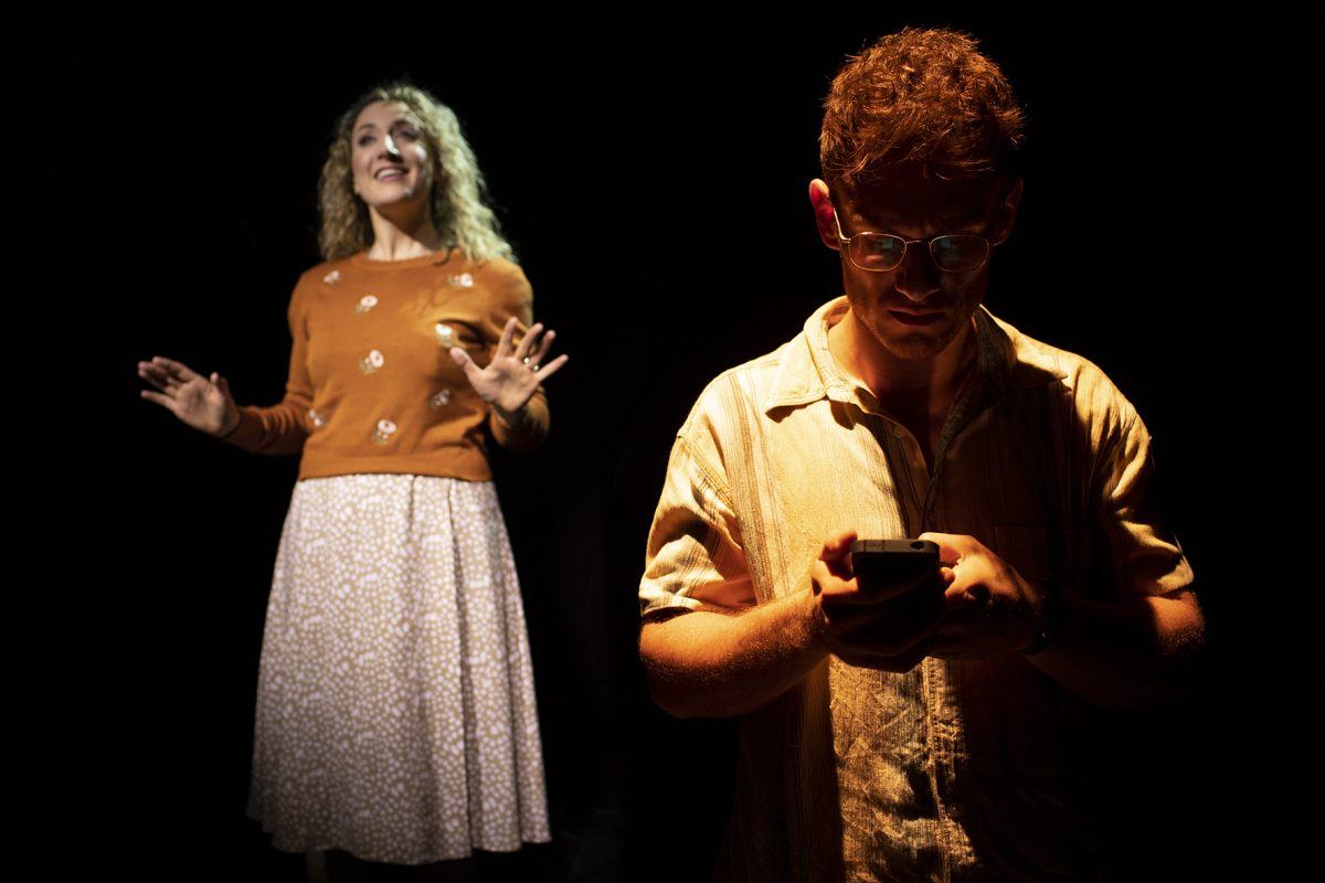 A woman in a floral skirt and orange jumper gestures expressively in soft light, whilst a man in glasses and a light shirt stands in the foreground, focused on his mobile, partially illuminated against a dark background.