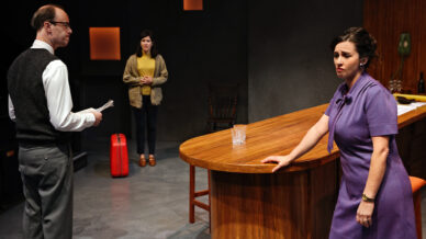 Three people are on a stage set. A man in glasses holds papers, a woman in purple leans on a bar looking concerned, and a woman with a red suitcase stands in the background. The setting looks modern and dramatic.