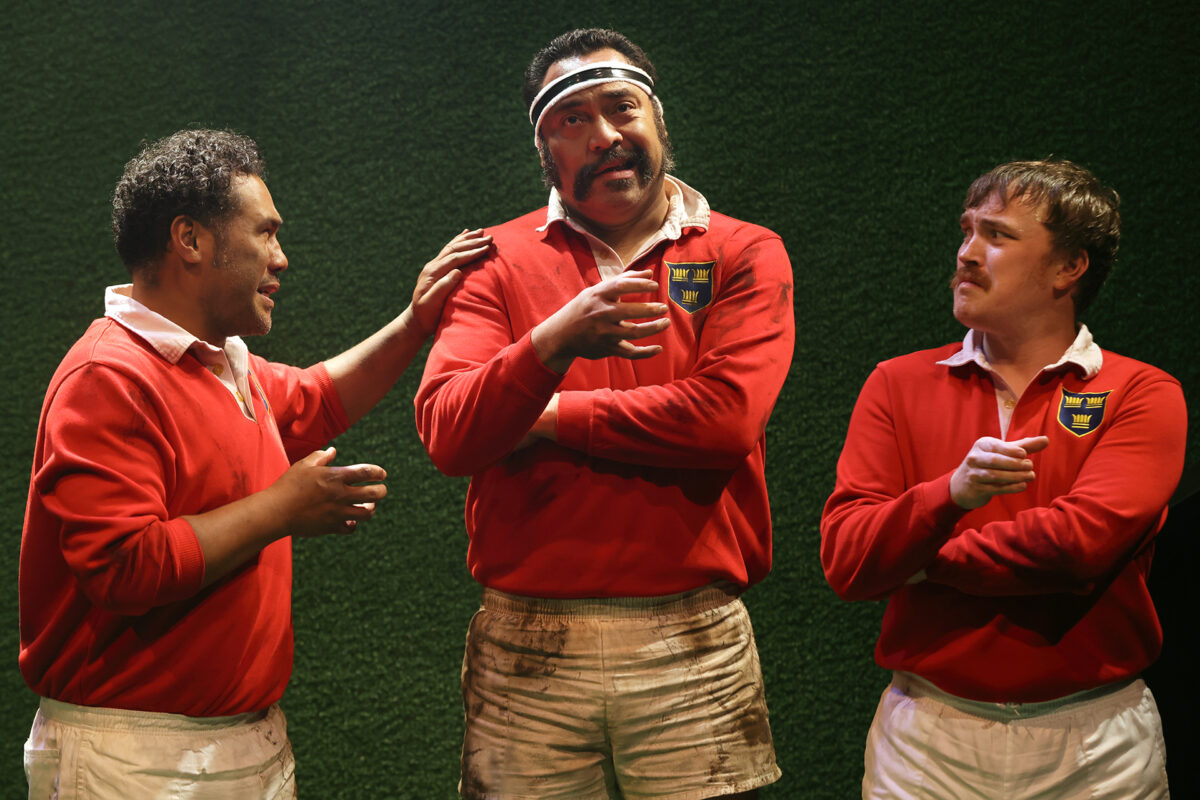 Three men in red rugby jerseys with white collars and muddy white shorts stand on a pitch. The man in the centre wears a headband and gestures while talking; the others listen and interact with him.