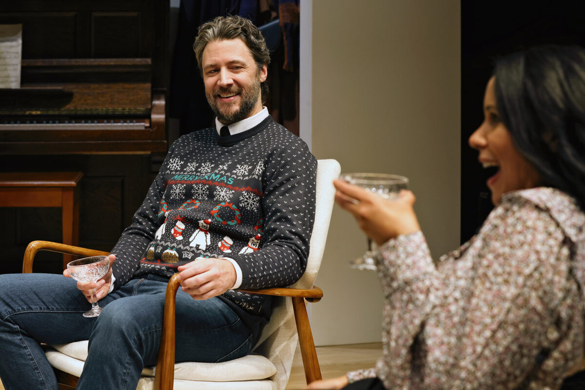 A man in a festive jumper sits smiling in a chair, holding a glass, while a woman beside him laughs and holds her own glass. A piano is visible in the background.
