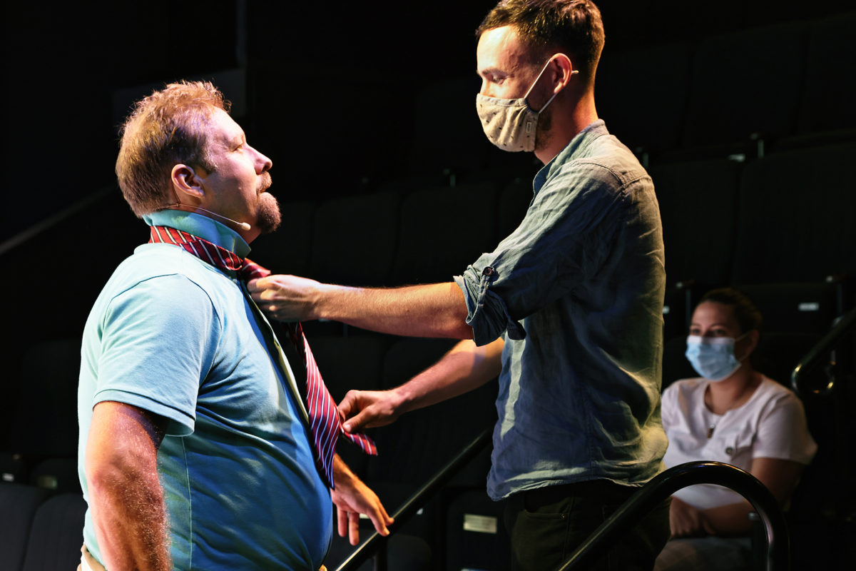 Two men wearing face masks stand indoors; one helps the other adjust his tie. A woman, also masked, sits in the background observing. The scene appears to be in a theatre or auditorium with empty seats.