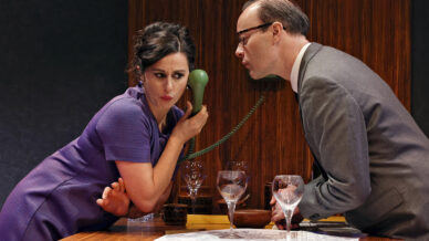 A woman in a purple dress listens intently to a green telephone whilst a man in a suit leans in, both looking serious. They are at a table with empty wine glasses and papers, against a wooden-panelled background.