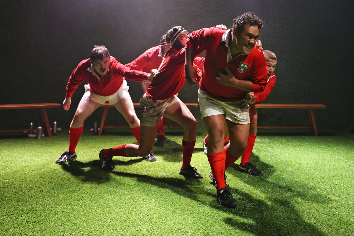 Four rugby players in red jerseys and white shorts run forward on a grassy pitch, with one player leading and others close behind. Their expressions are intense, and a wooden bench is visible in the background.