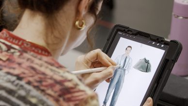 A person with curly hair uses a stylus to draw a fashion illustration of a person in a suit on a tablet. The tablet rests on a table alongside a purple water bottle and a black case.