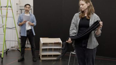 A woman stands holding a folded black umbrella, looking down, whilst a man in the background reads from a script. They are in a room with a black wall, a chair, a ladder, and a wooden structure.