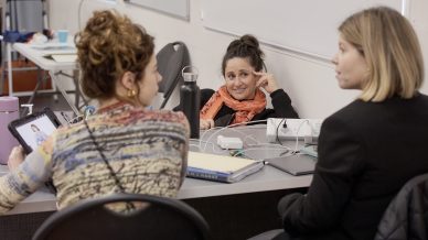 Three women sit around a table in an office or classroom, engaged in conversation. One woman faces the camera, smiling, while the other two sit with their backs or profiles towards the camera.