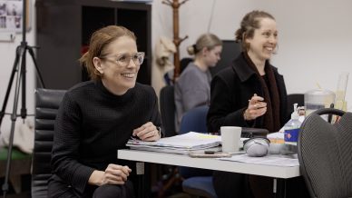 Two women sit at a desk, smiling and engaged in conversation. Papers, a mouse, and a coffee cup are on the table, with a third person working in the background in a bright office space.