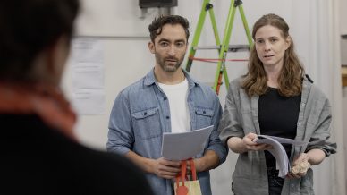 Two people holding scripts stand side by side, facing someone off camera. Both look attentive and engaged. A green ladder and sheets of paper hang on the wall in the background.