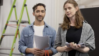 A man and woman stand indoors holding papers and looking ahead. A green ladder is visible in the background. The man wears a denim shirt over a white T-shirt, and the woman wears a black shirt with a grey overshirt.