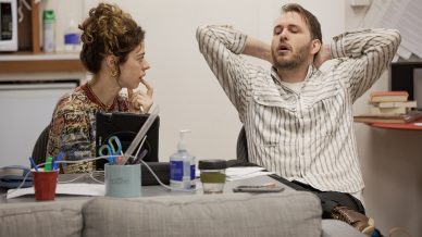 A woman sits talking to a man who is leaning back in his chair with his hands behind his head and eyes closed. A cluttered table with office supplies, hand sanitiser, and papers sits in front of them.