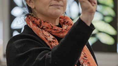 A woman with curly hair tied up, wearing an orange patterned scarf and black top, gestures with her hand whilst holding a book. She appears to be speaking or explaining something in an indoor setting. Leafy art décor is in the background.