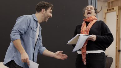 Two people rehearse a script together, laughing enthusiastically. One holds a notebook, and both appear animated and joyful, suggesting an engaging and lively moment during a rehearsal or acting session.