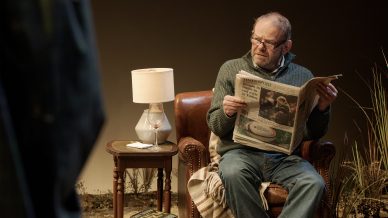 An older man sits in a brown leather armchair reading a newspaper. A lamp and a glass of water are on a small side table next to him, with some books stacked underneath. The setting appears cosy and warm.