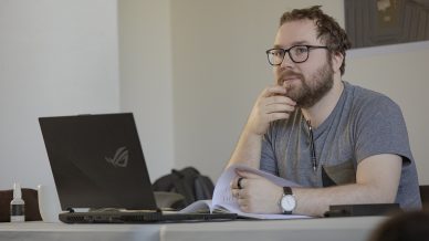 A bearded man with glasses sits at a table with a laptop, notebook, and pen, looking thoughtful. He wears a grey T-shirt and watch, with one hand touching his face and the other resting on an open book.