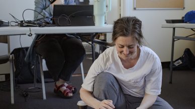 A woman sits on the floor barefoot, using a tablet to view a spiral-bound script. Behind her, another woman with headphones sits at a desk with a laptop, smiling. The room has plain walls and a noticeboard.