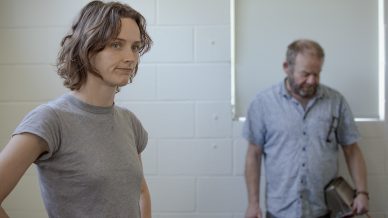 A young person with short, wavy hair and a grey t-shirt stands in focus in the foreground, looking to the side. In the background, an older man with a beard and blue shirt stands near a white brick wall, holding an object.