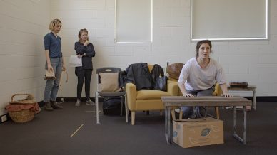 Three women in a rehearsal room; one sits on a box at a coffee table looking intense, while two others stand nearby watching her. The room has white walls, minimal furniture, and some props scattered about.