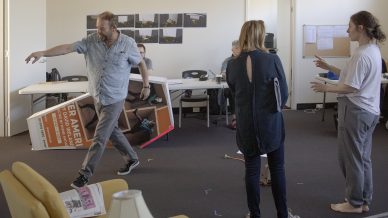 A man gestures animatedly while holding a large sign, surrounded by three people in a casual office setting with scattered papers and props. One person is barefoot, and others watch or interact with him.
