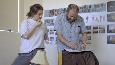 A woman stands on a side table next to a yellow armchair, watching a man write on a clipboard placed on the chair’s back. The room has photo collages on the wall, books, and everyday objects scattered around.