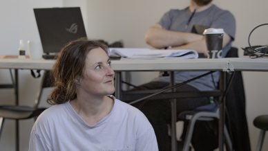 A woman in a white T-shirt and grey trousers sits cross-legged on the floor, looking to the side. Behind her, a man sits at a table with a laptop, papers, and a coffee cup, smiling and looking in the same direction.