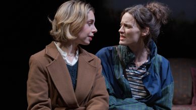 Two women sit closely on a sofa, facing each other and having an intense conversation. One wears a brown coat and has short blonde hair; the other has long brown hair tied up and is dressed more casually.