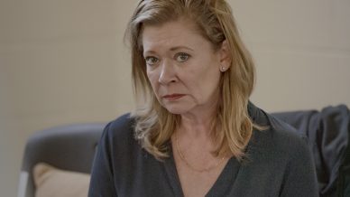 A woman with shoulder-length blonde hair sits on a yellow chair, holding an open folder with highlighted pages. She wears a dark blouse and looks concerned or worried. A sofa with cushions is in the background.